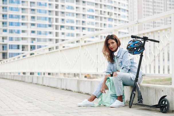 Mooie jonge Aziatische vrouw rusten op stoeprand na het rijden op elektrische scooter en het nemen van kopje van take-out koffie uit rugzak - Foto, afbeelding