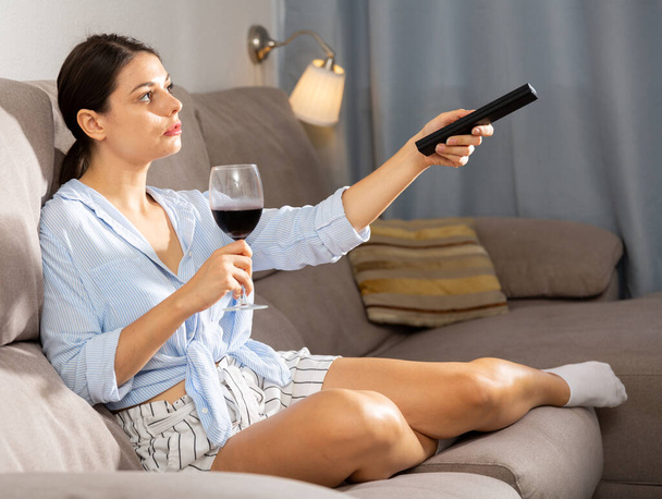 Młoda kobieta ze szklanką czerwonego wina ogląda telewizję w domu - Zdjęcie, obraz