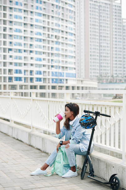 Ziemlich stylische junge Asiatin sitzt auf dem Bordstein, nachdem sie auf einem Elektroroller gefahren ist und Kaffee getrunken hat - Foto, Bild