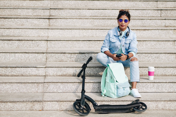 Güneş gözlüklü güzel bir kadın elektrikli scooter ve sırt çantasının yanındaki merdivenlerde dinleniyor mesaj atıyor ve kahve içiyor. - Fotoğraf, Görsel