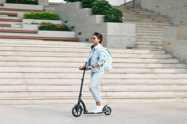 Üniversite kampüsünde elektrikli scooter kullanan, sırt çantalı güzel, şık genç bir kadın, bulanık hareket ediyor. - Fotoğraf, Görsel
