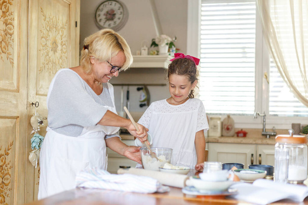 Babička a roztomilá malá kudrnatá vnučka v kuchyni, připravují se spolu. - Fotografie, Obrázek