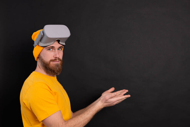 веселый красивый позитивный мужчина в очках VR указывает слева руками на черном фоне - Фото, изображение