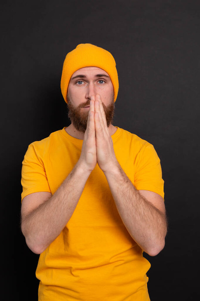 Portrait of bearded european man in yellow hat on black background praying with hands together asking for forgiveness smiling confident - Foto, Imagen