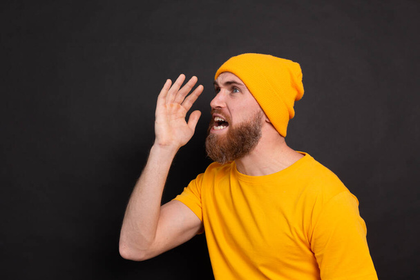 Portrait of bearded european man in yellow hat on black background shouting and screaming loud to side with hand on mouth. communication concept. - Foto, afbeelding