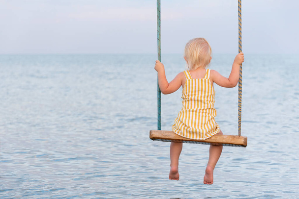 Blondynka w żółtej sukience siedząca na huśtawce liny nad wodą. Huśtawka po morzu. - Zdjęcie, obraz
