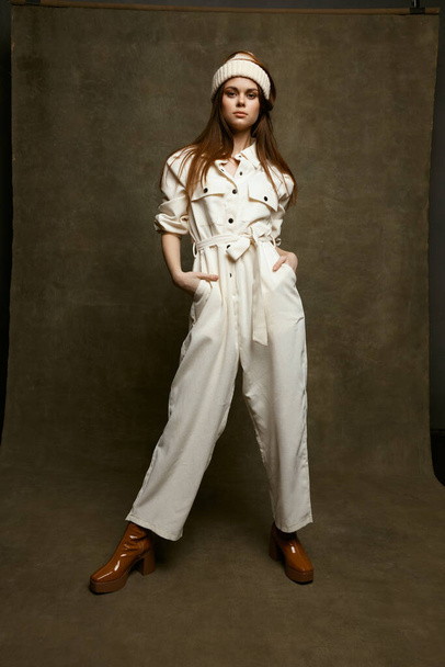 fashionable woman in a white jumpsuit holds hands in pockets and a hat on her head brown shoes - Foto, Imagen