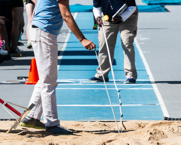 Funcionarios de pista que miden la distancia de un atleta saltan en el foso de arena con una pista azul al aire libre. - Foto, Imagen