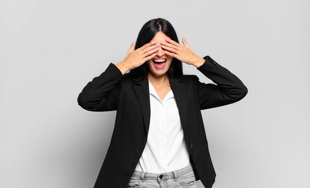 young hispanic businesswoman smiling and feeling happy, covering eyes with both hands and waiting for unbelievable surprise - Fotografie, Obrázek