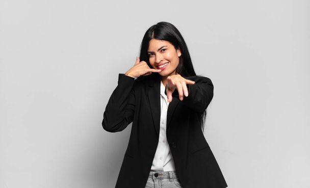 young hispanic businesswoman smiling cheerfully and pointing to camera while making a call you later gesture, talking on phone - Foto, Imagen