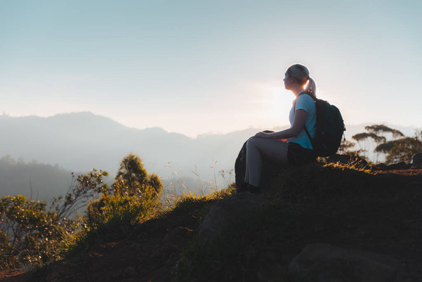 Sylwetka młodej podróżniczki cieszącej się spokojnym i inspirującym widokiem na zachód słońca lub wschód słońca po wycieczce na szczycie Little Adam 's Peak w górach Sri Lanki. - Zdjęcie, obraz