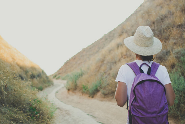 Rückansicht einer Wanderin, die auf einem Pfad in der Schlucht des Nationalparks wandert und einen lila Rucksack, einen sommerlichen Strohhut und ein weißes T-Shirt trägt. Aktives Reise- und Abenteuerkonzept. - Foto, Bild
