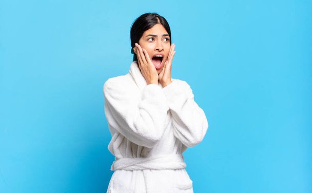 young hispanic woman feeling happy, excited and surprised, looking to the side with both hands on face. bathrobe concept - Φωτογραφία, εικόνα