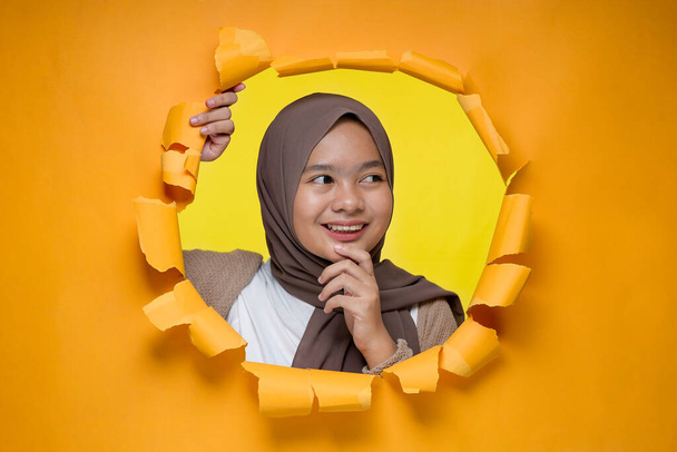 Młody azjatycki muzułmanin, uśmiechnięty nastolatek, pozuje przez rozerwaną żółtą dziurę w papierze, patrząc na przestrzeń kopiującą lub pusty żółty papier, nosząc hidżab, reklamując najlepszy produkt. Działalność reklamowa. - Zdjęcie, obraz