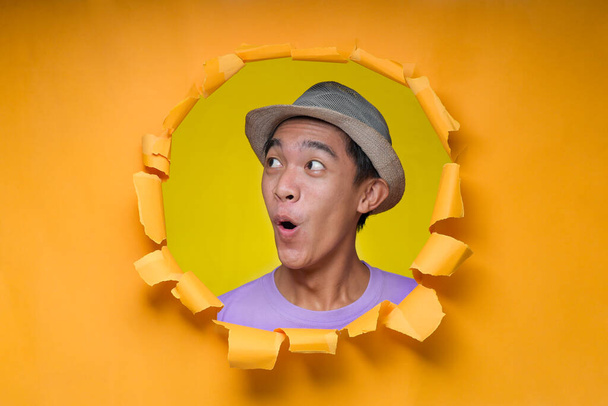 Hombre joven asiático con la cara impactada, expresión asombrada adolescente, posa a través de un agujero de papel amarillo roto, vistiendo una camiseta púrpura, asustado y sorprendido con la expresión sorpresa, el miedo y la cara emocionada - Foto, Imagen