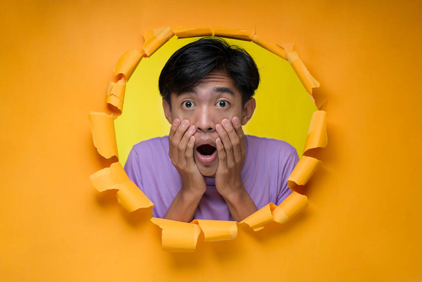 Azji Młody człowiek z wstrząśnięty, zdumiony wyraz twarzy nastolatek, pozuje przez rozdarty żółty otwór papieru, w fioletowy t-shirt, boi się ekspresji - Zdjęcie, obraz