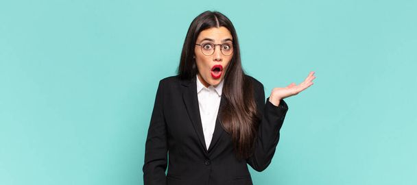 young pretty woman looking surprised and shocked, with jaw dropped holding an object with an open hand on the side. business concept - Foto, Bild