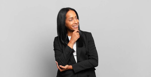 young black woman smiling with a happy, confident expression with hand on chin, wondering and looking to the side. business concept - Foto, imagen
