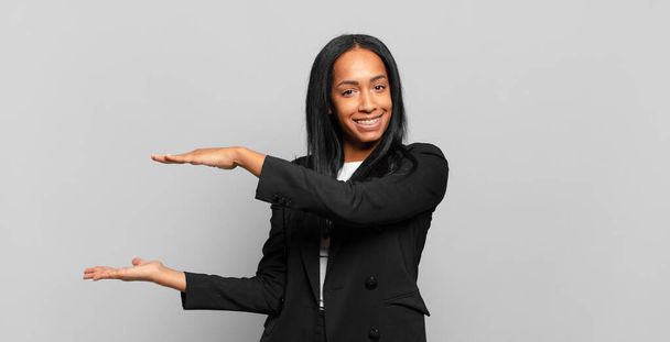 young black woman smiling, feeling happy, positive and satisfied, holding or showing object or concept on copy space. business concept - Foto, imagen