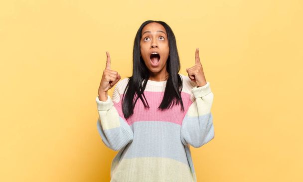young black woman looking shocked, amazed and open mouthed, pointing upwards with both hands to copy space - Photo, image