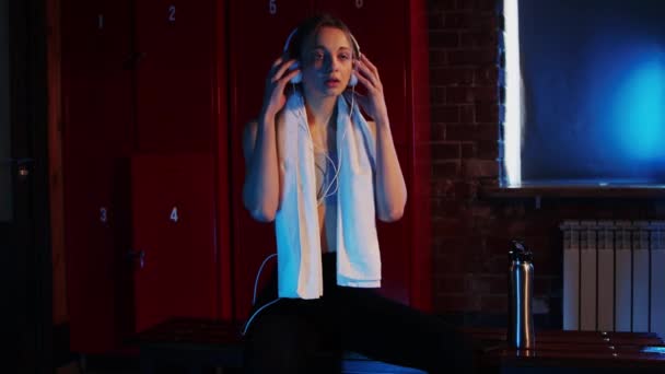 Sport - fiatal csinos nő ül az öltözőben egy törülköző és fejhallgató a nyakán - Felvétel, videó