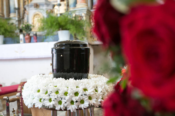 Schwarze Urne am Tag der Beerdigung in der Kirche; umgeben von Gänseblümchen und Rosen - Foto, Bild