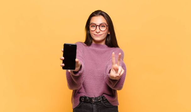 young pretty woman smiling and looking happy, carefree and positive, gesturing victory or peace with one hand. phone screen concept - Foto, Imagem