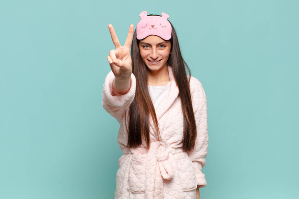 young pretty woman smiling and looking happy, carefree and positive, gesturing victory or peace with one hand. awaking wearing pajamas concept - Φωτογραφία, εικόνα