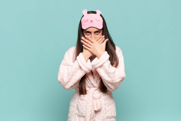 young pretty woman covering mouth with hands with a shocked, surprised expression, keeping a secret or saying oops. awaking wearing pajamas concept - Foto, imagen
