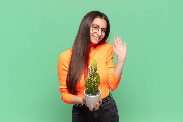 young pretty woman smiling happily and cheerfully, waving hand, welcoming and greeting you, or saying goodbye. cactus concept - Fotografie, Obrázek