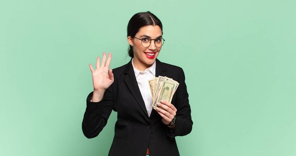 young pretty woman smiling happily and cheerfully, waving hand, welcoming and greeting you, or saying goodbye. business and banknotes concept - 写真・画像