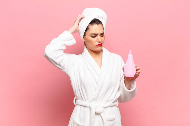 young pretty woman feeling puzzled and confused, scratching head and looking to the side. bathrobe and face cleaning concept - Photo, image