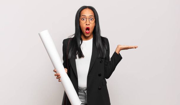 young black woman looking surprised and shocked, with jaw dropped holding an object with an open hand on the side. architect concept - Photo, image