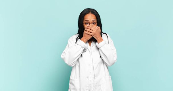 junge schwarze Frau bedeckt Mund mit Händen mit einem schockierten, überraschten Gesichtsausdruck, hält ein Geheimnis oder sagt uups. Ärztekonzept - Foto, Bild