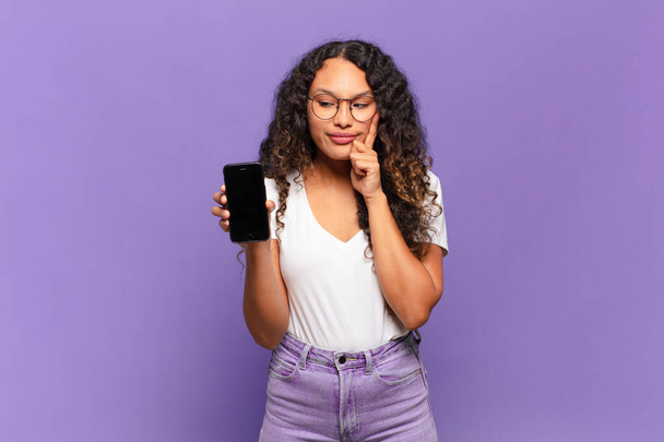 young hispanic woman smiling with a happy, confident expression with hand on chin, wondering and looking to the side. smart phone concept - Φωτογραφία, εικόνα