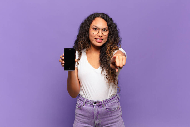 young hispanic woman pointing at camera with a satisfied, confident, friendly smile, choosing you. smart phone concept - Фото, изображение