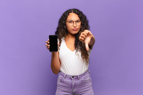 young hispanic woman feeling cross, angry, annoyed, disappointed or displeased, showing thumbs down with a serious look. smart phone concept - Fotoğraf, Görsel