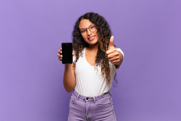 young hispanic woman feeling proud, carefree, confident and happy, smiling positively with thumbs up. smart phone concept - Photo, Image