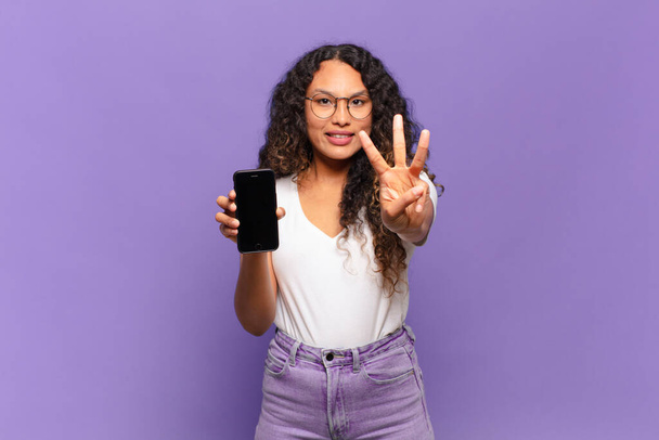 young hispanic woman smiling and looking friendly, showing number three or third with hand forward, counting down. smart phone concept - Φωτογραφία, εικόνα