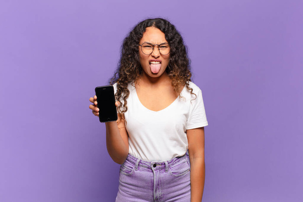 young hispanic woman feeling disgusted and irritated, sticking tongue out, disliking something nasty and yucky. smart phone concept - Foto, Imagen