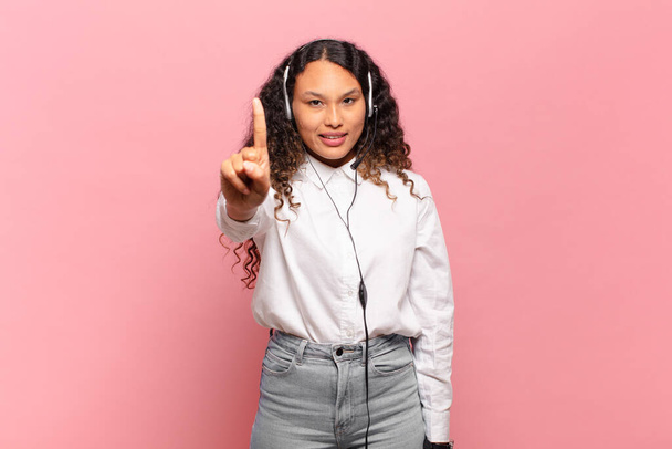 young hispanic woman smiling and looking friendly, showing number one or first with hand forward, counting down. telemarketer concept - Foto, Bild