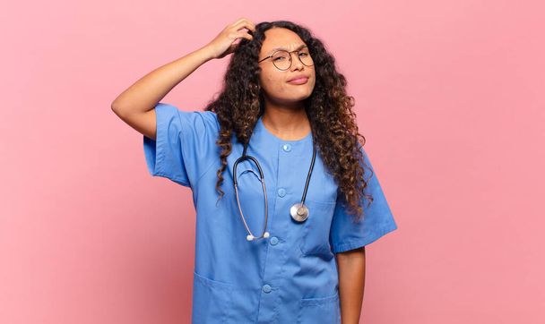 young hispanic woman feeling puzzled and confused, scratching head and looking to the side. nurse concept - Photo, Image
