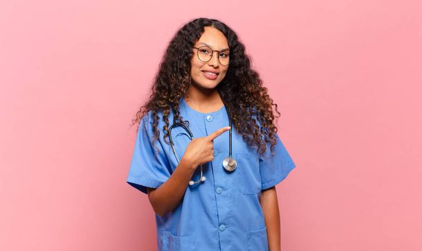 young hispanic woman smiling cheerfully, feeling happy and pointing to the side and upwards, showing object in copy space. nurse concept - Фото, изображение