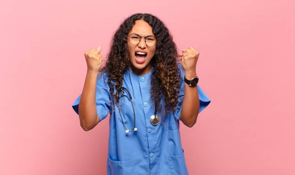 young hispanic woman shouting aggressively with an angry expression or with fists clenched celebrating success. nurse concept - Foto, Bild