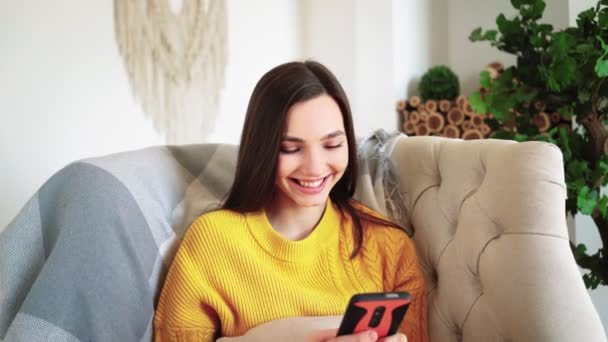 Mujer sonriente Hold Smartphone Ver historias de redes sociales Video Siéntate en el sofá - Metraje, vídeo