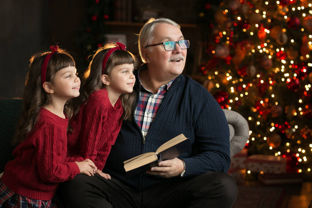 Grandfather wearing glasses reading a book to small granddaughters twins in a room decorated for Christmas amid a Christmas tree staring out the window. Christmas holiday concept. bright contrast photography - Photo, image
