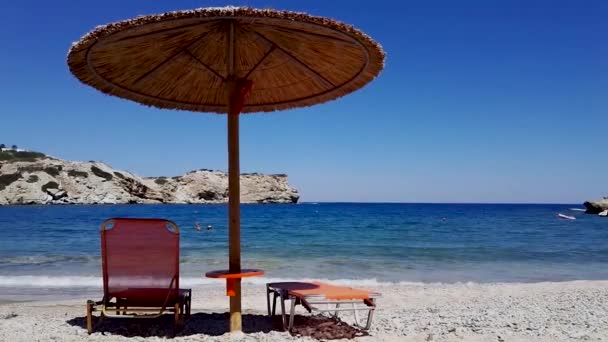 Foto estática de una sombrilla de hierba y banco de sol en la arena contra el mar azul mediterráneo de la playa de Herkalion contra la isla rocosa en Creta, Grecia. Tiro estático de banco contra las olas del mar. - Imágenes, Vídeo