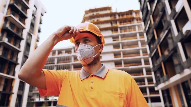 El tema es la protección de la salud en la producción y construcción. Caucásico trabajador masculino en uniforme naranja y protector respirador kn 95 pone en la construcción naranja sombrero duro y posa en el sitio de construcción. - Foto, imagen