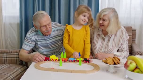 Child girl kid involved in build board game ride toy train on railroad with senior grandparents home - Footage, Video