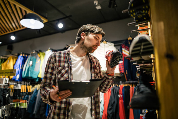 Business owner in sports store works with showcase trekking and mountaineering boots for sale, holding clipboard in his hands, checks availability of goods and collects to process online orders. - Photo, Image